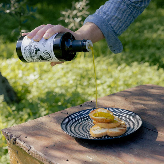 Honest Toil Olive Oil 500ml being poured over a plate of food in a natural outdoor setting.