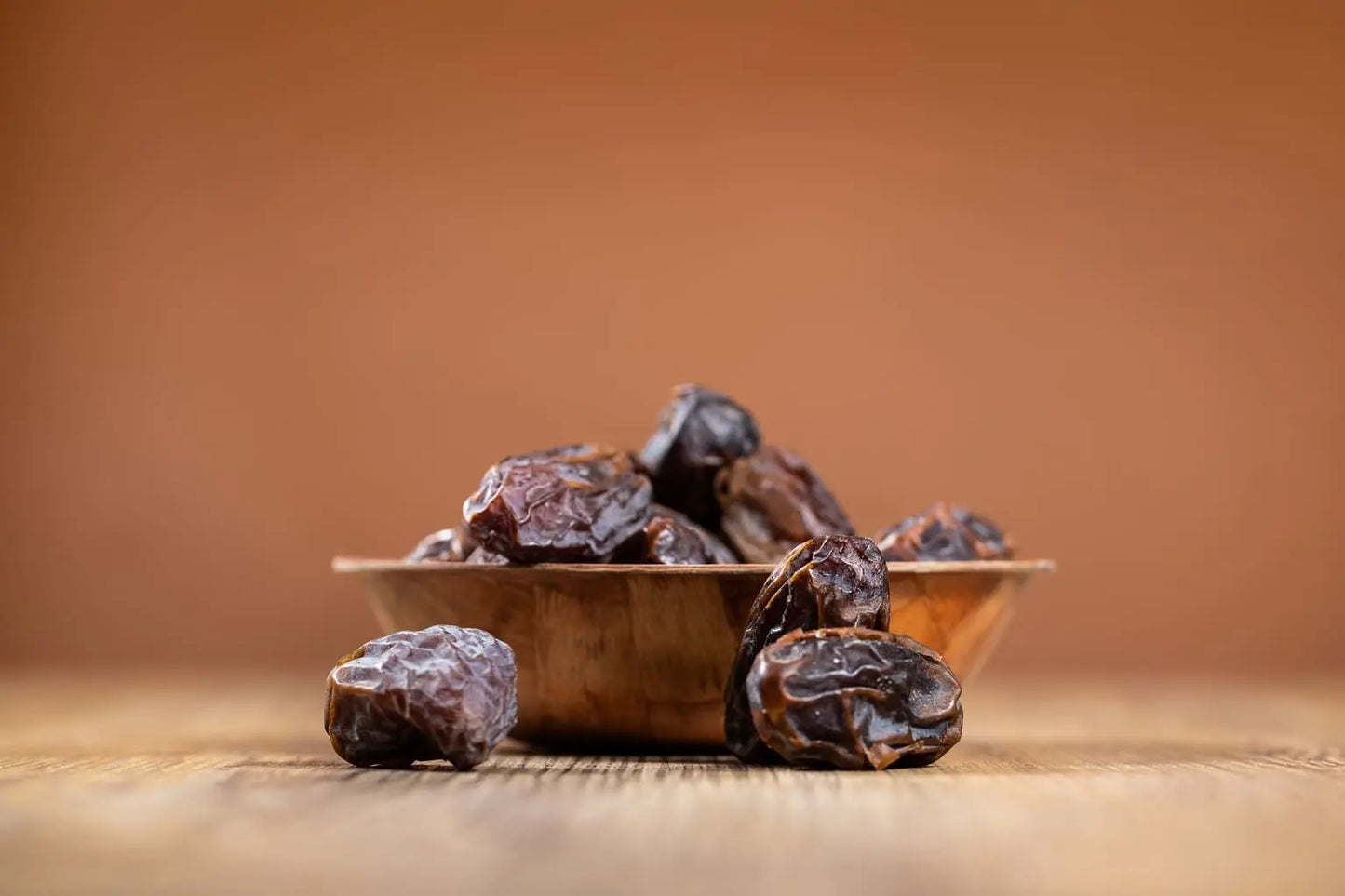 Several dark brown dates piled in a small wooden bowl on a wooden surface, with a warm brown blurred background.