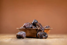 Several dark brown dates piled in a small wooden bowl on a wooden surface, with a warm brown blurred background.