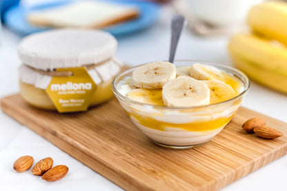 Mellona Raw Honey 230g jar next to a bowl of yogurt topped with banana slices and almonds on a wooden surface.